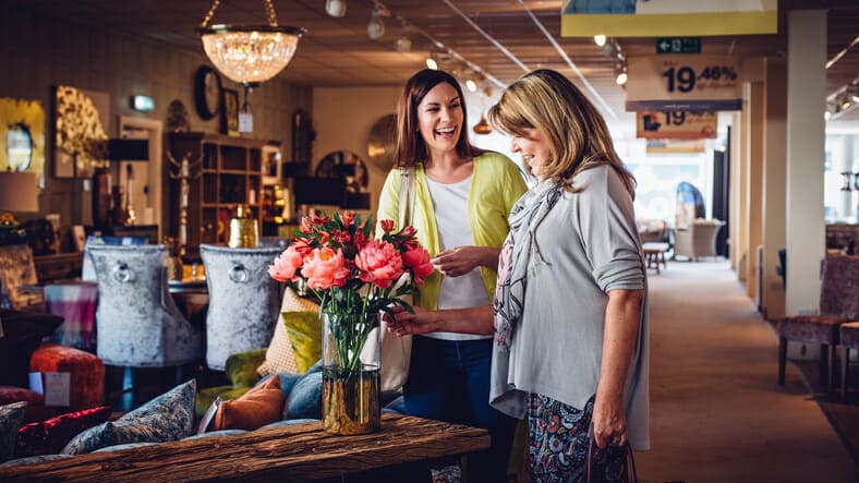 Females working in the retail business. They stand beside a vase of flowers and smiling standing in a large furniture store. Home furniture can be seen around them as they arrange and plan together.